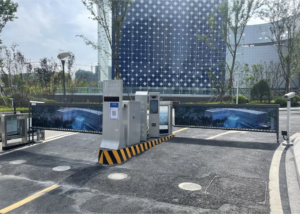 A modern parking lot entrance with automated ticket machines and barriers, set against a backdrop of contemporary buildings with glass facades and trees lining the area.